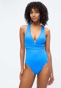 Γυναίκα με κυματιστά μαλλιά, φορούν λαμπερό μπλε ολόσωμο μαγιό με βαθύ V στο λαιμό και σούρα στη μέση, στέκεται μπροστά σε απλό φωτεινό φόντο.