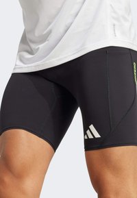 Schwarze Sportshorts mit enger Passform, ausgestattet mit einem weißen Adidas-Logo, grünen Akzenten und flachen Nähten für Komfort und Flexibilität.