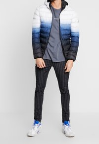 Gevoerde jas in wit, blauw en zwart verloop. Voorzien van een ritssluiting aan de voorkant en een capuchon. Gekleed met een grijs t-shirt en gestreepte zwarte broek.