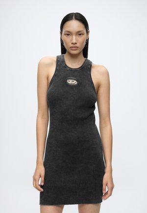 Femme aux cheveux noirs et lisses porte une robe sans manches côtelée gris foncé avec une petite découpe ovale près de la clavicule et des boucles d’oreilles créoles argentées.
