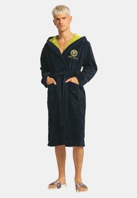 Marineblauwe pluche badjas met een groene, gedessineerde binnenvoering van de capuchon, een taille-touw, zakken en geborduurd logo. Contraststof rondom de capuchon.
