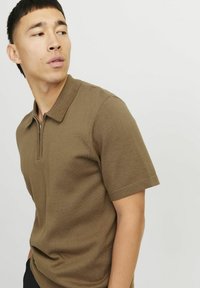 Polo en khaki avec un col zippé, des manches courtes et un tissu texturé. Le vêtement a une coupe décontractée et des éléments de design minimalistes.