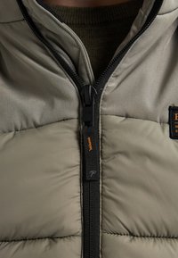 Giacca khaki con zip, caratterizzata da un design imbottito, zip nera con un accento arancione e un tessuto testurizzato. Cuciture visibili lungo le cuciture.