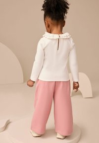 Bambino con capelli ricci in piedi di spalle, indossa una maglietta bianca a maniche lunghe con colletto arricciato e pantaloni rosa a gamba larga.