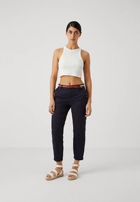 Ženska stoji oblečena v belo kratko majico brez rokavov, temno modre hlače z rjavim pasom in bele sandale z naramnicami pred enobarvno ozadje.