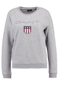 Sudadera gris con cuello redondo, mangas raglán y logo de GANT. Presenta un parche en forma de escudo con acentos de color rojo, blanco y azul.