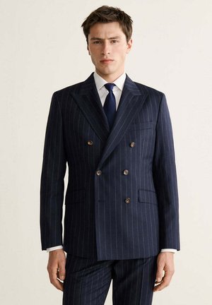 Hombre con traje azul marino a rayas de doble botonadura, camisa blanca y corbata oscura, de pie frente a un fondo claro y sencillo.