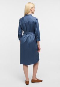 Satinblaues Kleid mit Kragen, Dreiviertelärmeln, Krawattengürtel in der Taille, rückseitigen Falten und seitlichen Schlitzen für Bewegungsfreiheit.