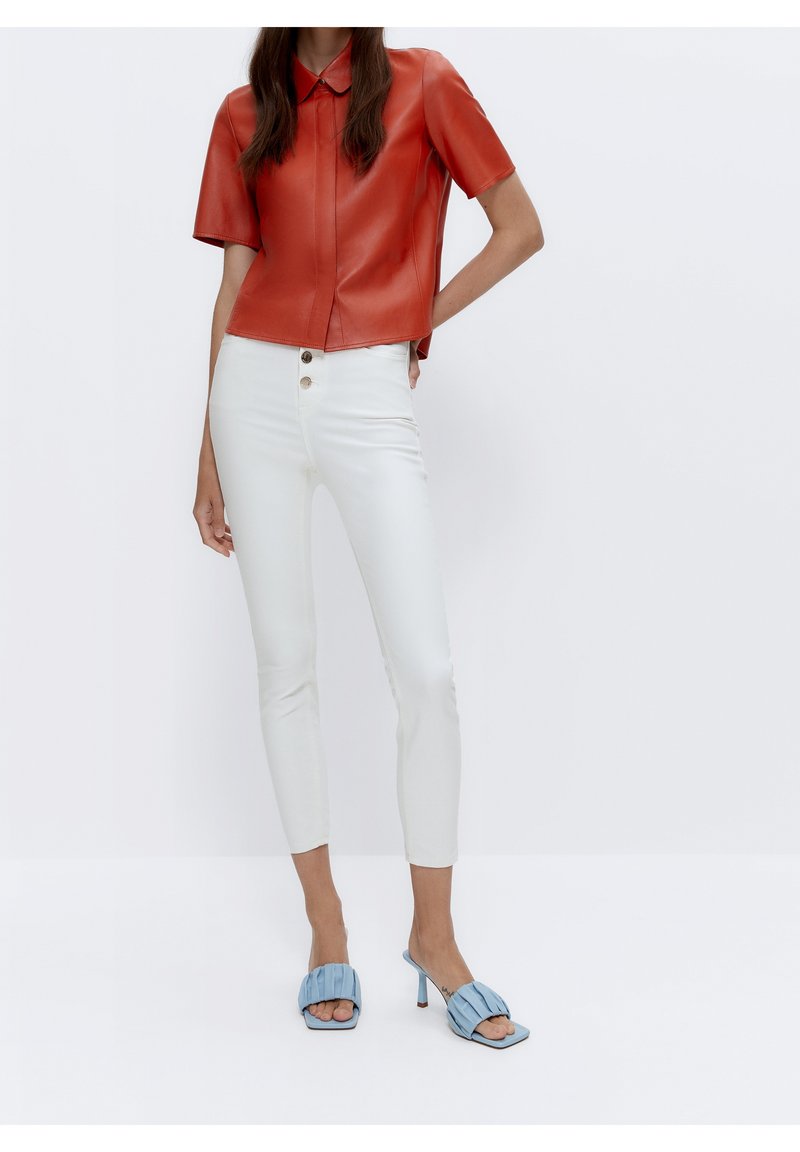 Camisa corta de cuero rojo con cuello, combinada con pantalones ajustados blancos y sandalias de tacón azules con diseño plisado.