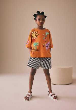 Ung pige står med hænderne i lommerne iført en orange skjorte med sol, palme og sommerfugleprint, sort-hvide ternede shorts og hvide sandaler.
