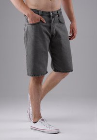 Male wearing gray knee-length denim shorts and white sneakers, standing with one hand in pocket against plain gray background.