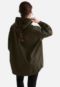 Femme aux cheveux tressés ajustant la capuche d'une veste de pluie vert foncé surdimensionnée, debout devant un fond blanc uni.