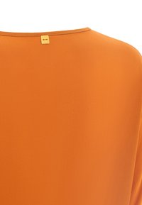 Orange Bluse mit glatter Stoffoberfläche, runder Ausschnitt und kleinem goldenen Etikett auf der Rückseite. Einfaches und schmuckloses Design.