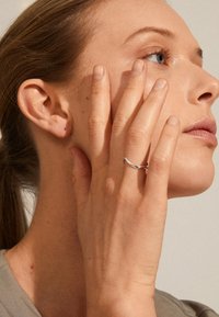 Anello a forma di onda in argento su una mano che tocca la guancia; primo piano sulla texture della pelle con trucco leggero e naturale, su uno sfondo neutro.