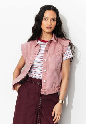 Femme aux longs cheveux foncés porte une veste en jean rose sans manches, un t-shirt rayé blanc et rouge, un pantalon bordeaux et un bracelet doré, debout contre un mur blanc.