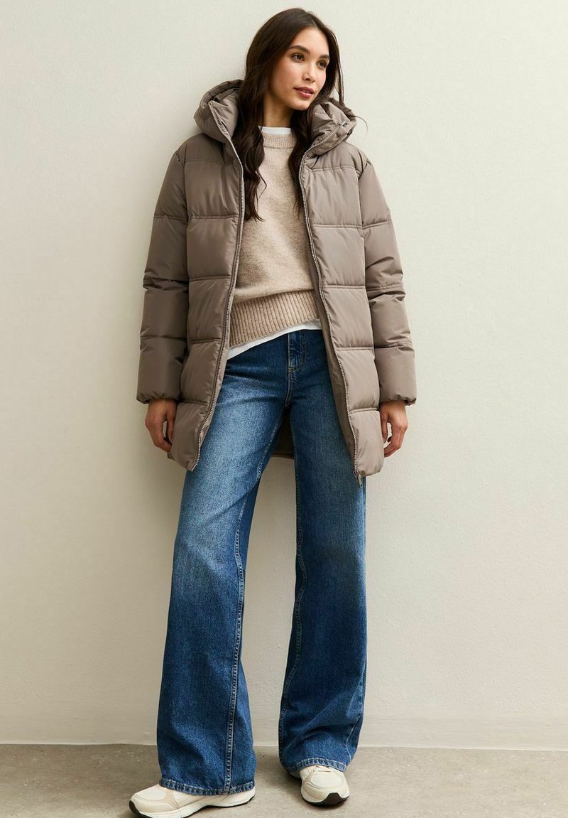 Vrouw staat in een taupe pufferjack, beige sweater, wijde blauwe jeans en witte sneakers tegen een effen lichte muur.