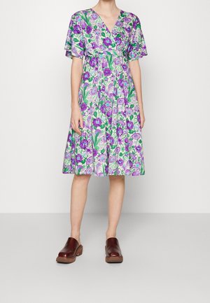 Person iført en knælængde lilla og grøn blomsterkjole med korte ærmer og brune slip-on sko, stående foran en ensfarvet lys baggrund.