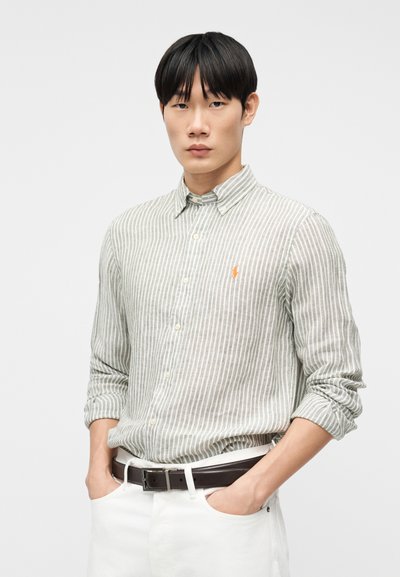 Νεαρός άνδρας που φοράει πουκάμισο με κουμπιά σε ανοιχτό γκρι και λευκές ρίγες με τα μανίκια γυρισμένα, λευκό παντελόνι και σκουρό καφέ ζώνη, στέκεται με τα χέρια στις τσέπες.