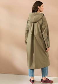 Olivegrüner langer Mantel mit Kapuze, geraffter Taille und verstellbaren Manschetten. Kombiniert mit hellblauen, cropped Jeans und burgunderfarbenen Sneakers.