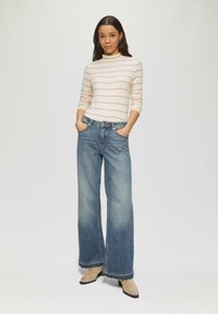 Strickpullover mit hellrosa Streifen, kombiniert mit weit geschnittenen blauen Jeans. Die Jeans haben einen fransen Saum und Taschen; das Model steht mit den Händen in den Taschen.