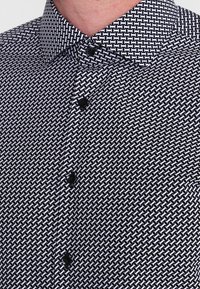 Lyhythihainen paita, jossa mustavalkoinen geometrinen kuviointi, nappikiinnitteinen kaulus ja mustat napit. Kangas näyttää sileältä ja kevyeltä.