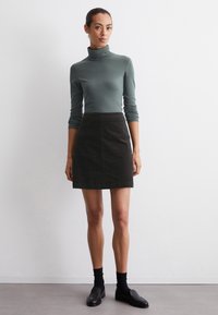 Een langslavend, aansluitend coltrui in een gedempte groene stof, gecombineerd met een zwarte A-lijn minirok, gedragen met zwarte loafers en enkelsokken.