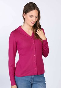 Langärmliges fuchsia Shirt mit Kragen, ausgestattet mit einer durchgehenden Knopfleiste und einer figurbetonten Passform, getragen über blauen Jeans. Glatte Textur, unifarbenes Muster.