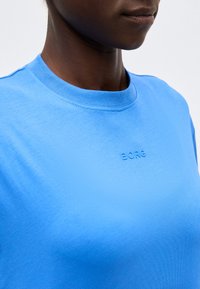 Κοντινό πλάνο ενός ατόμου που φοράει φωτεινό μπλε μπλουζάκι με στρογγυλή λαιμόκοψη με τη λέξη «BORG» ανάγλυφη στο στήθος.