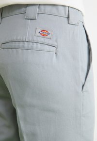 Pantaloni din bumbac gri cu o textură netedă, având un buzunar la spate și o etichetă țesută cu un logo circular. Cotele întărite sunt vizibile.