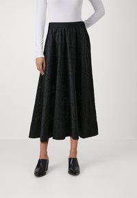 Falda maxi negra con patrón de ojales, tela texturizada y cintura elástica. Combinada con mulas negras con tacón cuadrado.