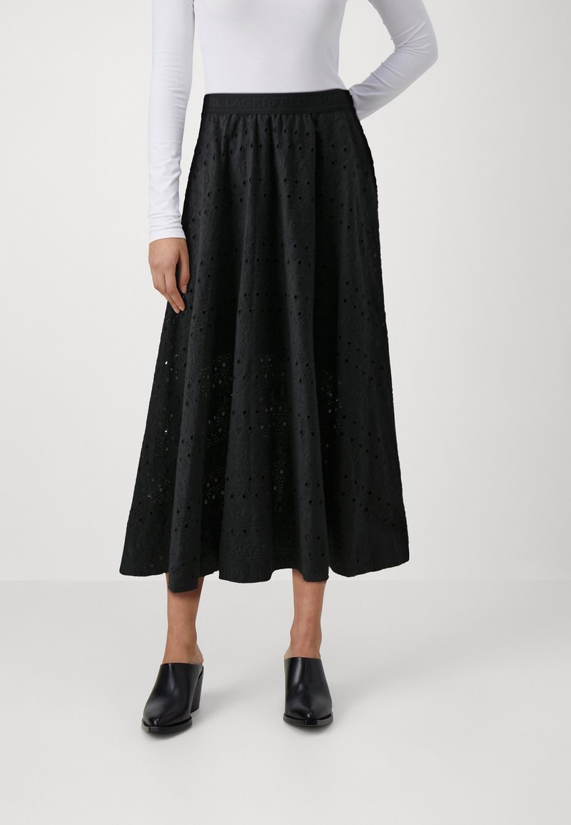Falda maxi negra con patrón de ojales, tela texturizada y cintura elástica. Combinada con mulas negras con tacón cuadrado.