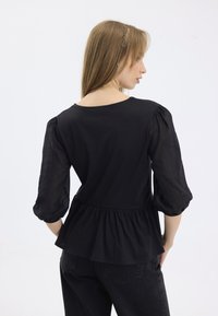 Svart peplum-top med trekvart lange gjennomsiktige ermer og rund hals. Jevnt stoff med detalj av rynket midje.