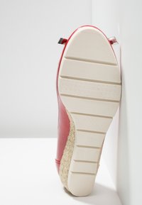 Chaussure en cuir rouge à surface texturée, semelle en caoutchouc blanc et détails en jute tissé autour de la base. Comprend un accent métallique sur le talon.
