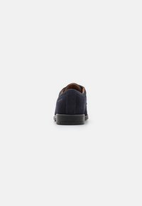 Zapato de ante azul marino con suela de goma negra, que presenta una textura suave y un diseño minimalista. Acentos marrones visibles alrededor del cuello del tobillo.