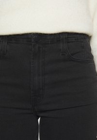 Jeans de mezclilla de cintura alta negra con cierre de botón, bolsillos frontales visibles y detalles de costura sutiles. Textura suave y forma ajustada.