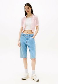 Top corto de crochet rosa claro con botones sobre una camiseta blanca ajustada, combinado con pantalones cortos bermuda de mezclilla azul claro y zapatillas blancas.