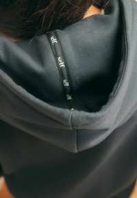 Donkergrijze hoodie met een zachte textuur, voorzien van een aantrekkoord in de capuchon en een wit "self." label genaaid nabij de halslijn.