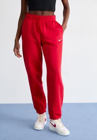 Červené Nike tepláky s elastickým pásom a šnúrkou, s bočnými vreckami a rebrovanými manžetami. Spárované s bielymi teniskami.