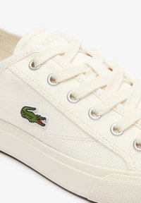 Gros plan sur une basket blanc cassé avec des lacets blancs, des œillets métalliques et un logo crocodile vert brodé sur le côté.