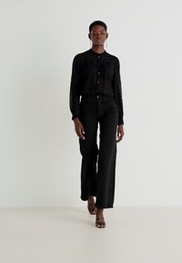 Chemisier noir en dentelle à manches longues avec fermeture à boutons, associé à un pantalon évasé noir taille haute. Le mannequin porte des talons noirs à lanières.