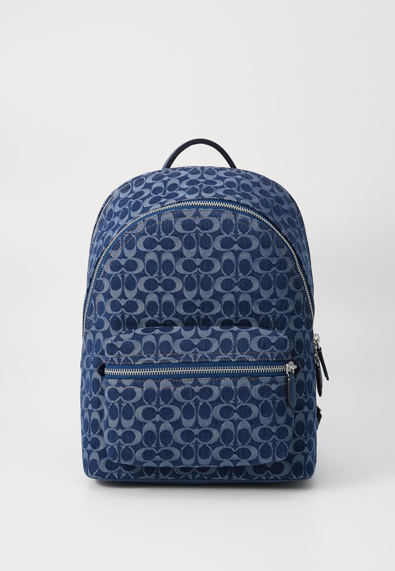 Sac à dos en denim bleu avec motif circulaire, poche frontale zippée et poignée supérieure. Texture lisse et détails de couture renforcée.
