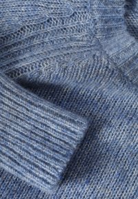 Pull bleu tricoté, présentant un tissu texturé avec un motif côtelé. Le matériau semble doux et duveteux, soulignant une apparence chaude.