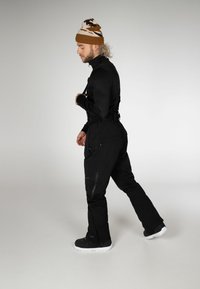 Pantaloni da neve neri con bretelle regolabili, numerose tasche con zip e design a gamba affusolata, abbinati a un berretto a fantasia marrone e bianco.