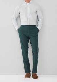 Chemise à rayures bleu clair et blanc avec boutons, associée à un pantalon chino vert foncé et des chaussures en daim marron. Design minimaliste, coupe ajustée.