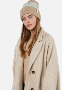 Beige knitted beanie with a blue and white geometric pattern, featuring a pom-pom on top. Soft texture and ribbed design for warmth.