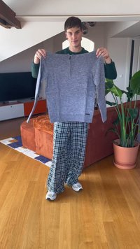 Young person indoors holding up a grey knitted sweater, wearing green plaid pants and white sneakers, with orange couch and plants in background.