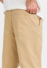 Beige pants made of woven fabric, featuring two back pockets, a side pocket, and a small brand label on the waistband.