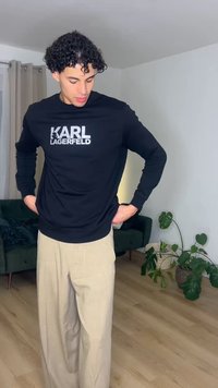 Noor mees lokkis juustega, kandes musta Karl Lagerfeldi pusa ja beeže pükse, seisab toas, kus on taimi ja roheline diivan.