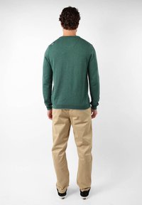 Pull en maille verte à manches longues et col rond, avec un détail de bouton sur l'épaule, porté avec un pantalon beige et des chaussures noires.