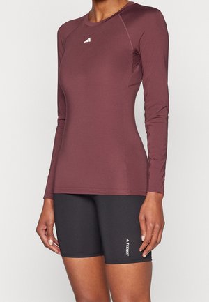 Frau trägt ein eng anliegendes, langärmeliges Sportoberteil in Kastanienbraun und schwarze Sportshorts bis zur Mitte des Oberschenkels mit "TECHFIT"-Logo.